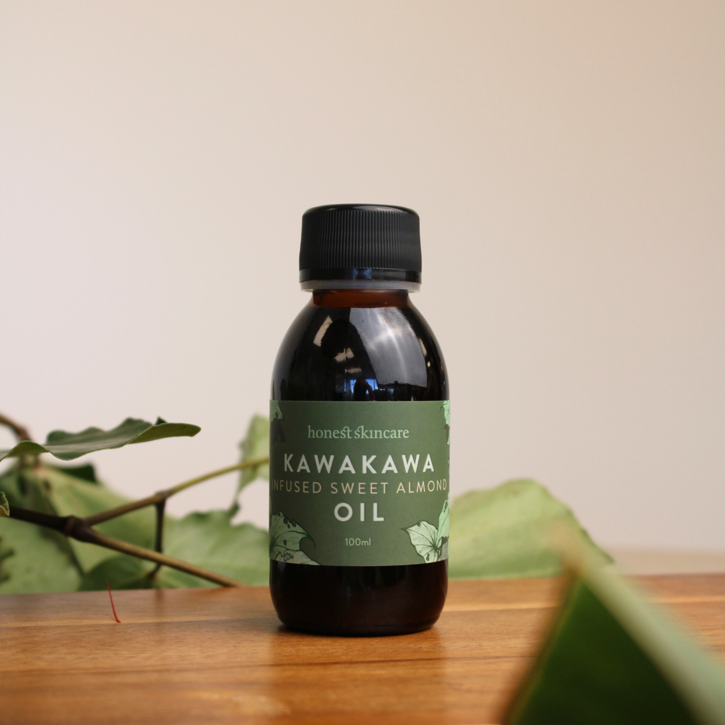 Bottle of Kawakawa infused sweet almond oil on a wooden surface with green leaves in the background.