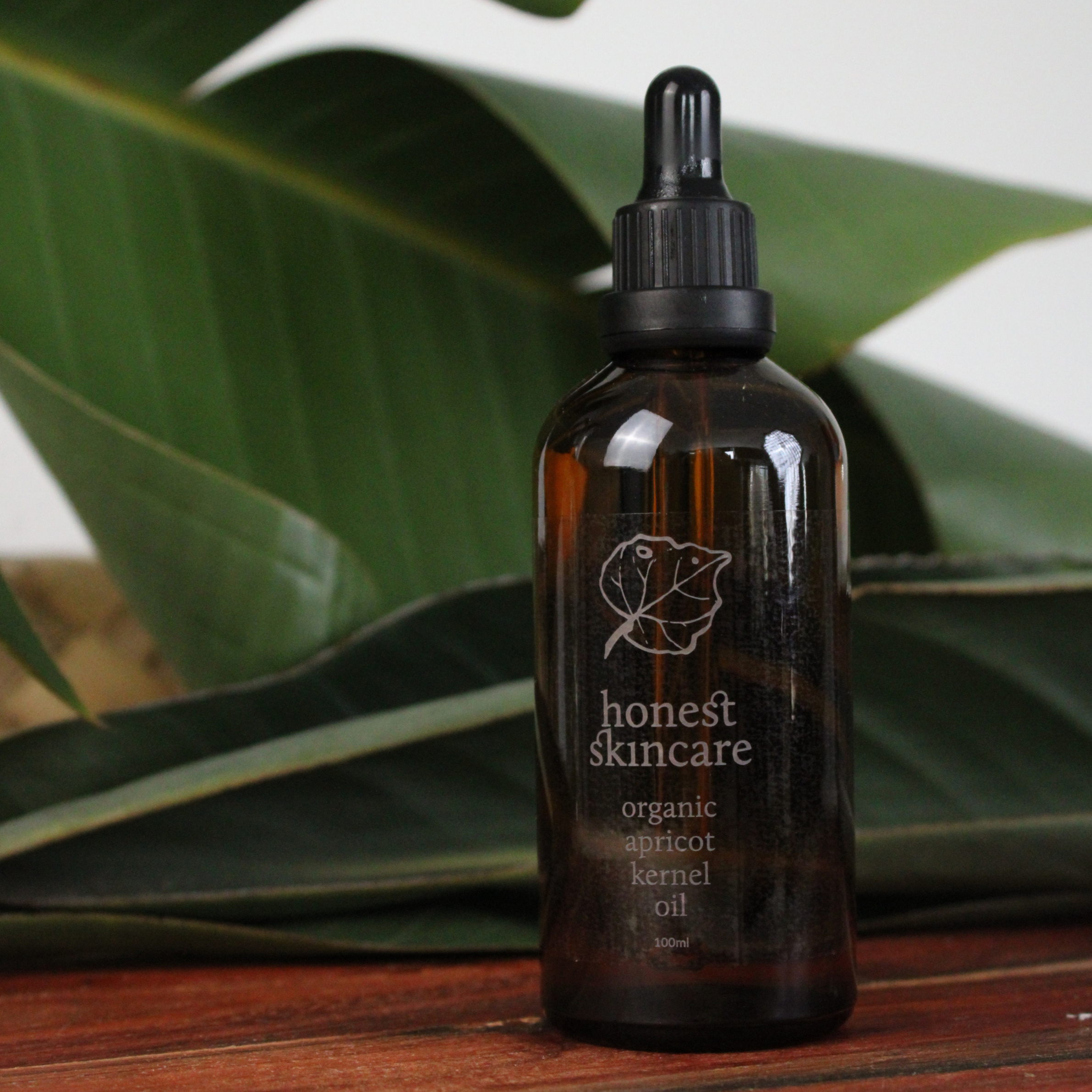 Brown glass bottle with black dropper labeled 'apricot kernel oil 100ml honest skincare' on a wooden surface with green leaves in the background.