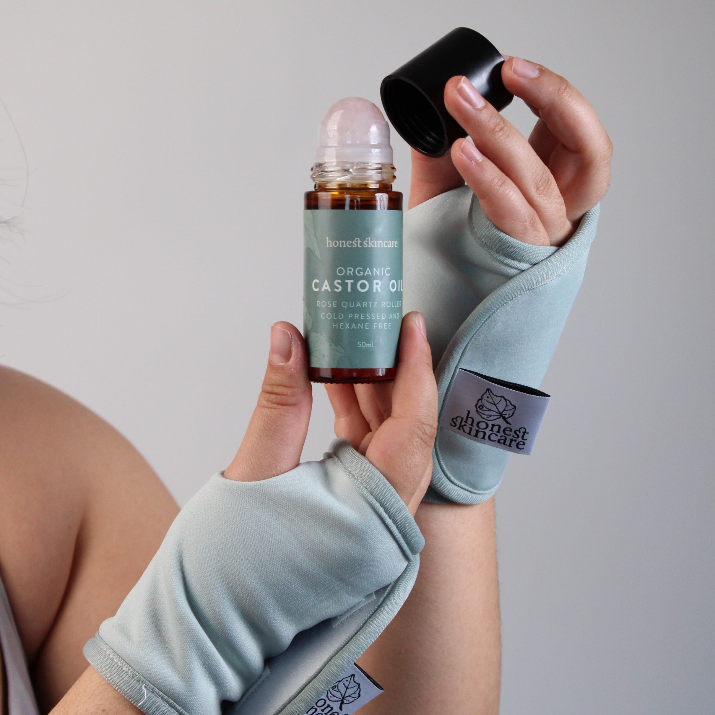 A person wearing light blue fingerless wrist wraps holds a roll-on bottle of castor oil with its cap off, set against a plain light gray background.