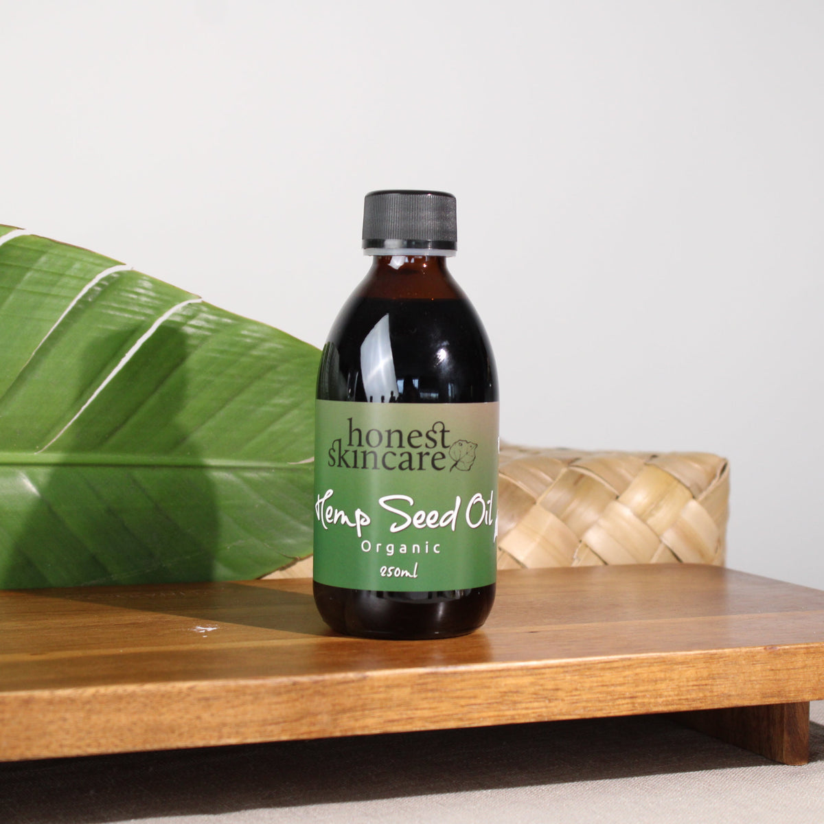 A bottle of Honest Skincare Hemp Seed Oil stands on a wooden tray, with a large green banana leaf on the left and a woven basket in the background against a light gray wall.