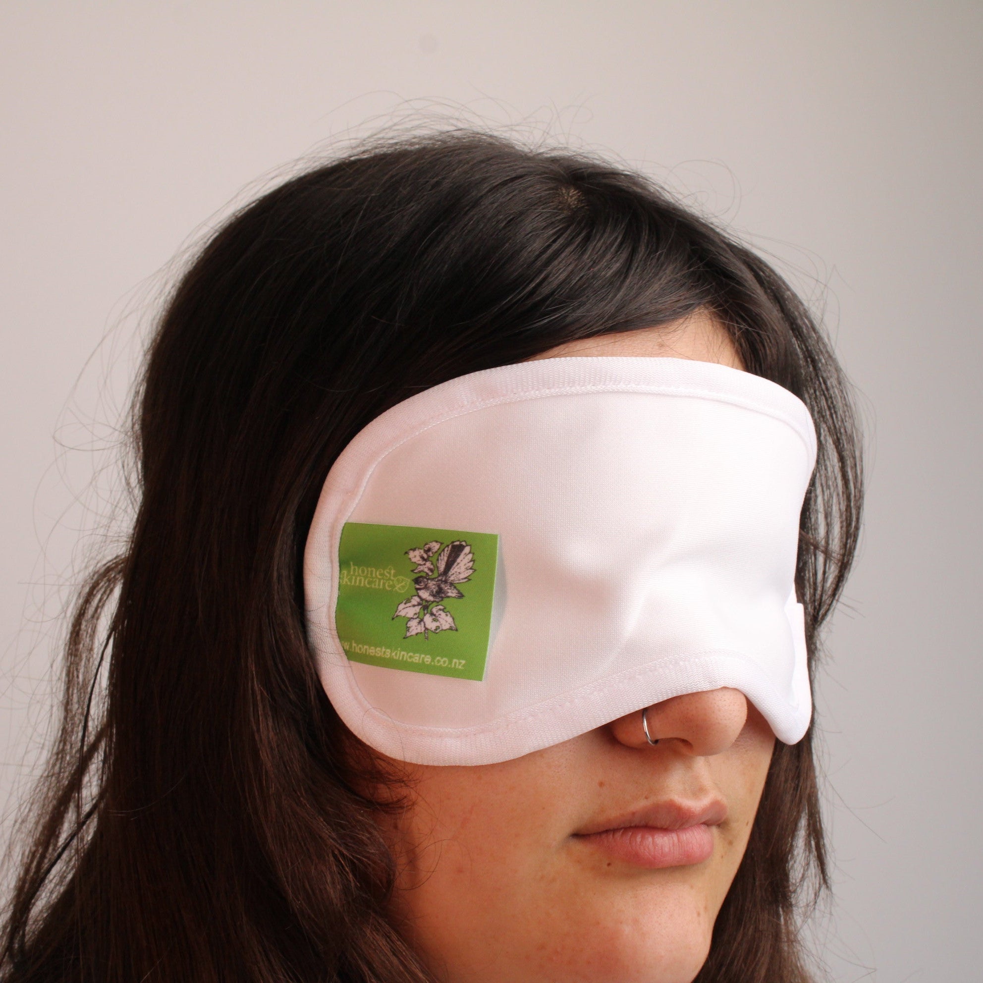 Person wearing a white organic cotton castor oil eye mask with a green honest skincare label against a plain background