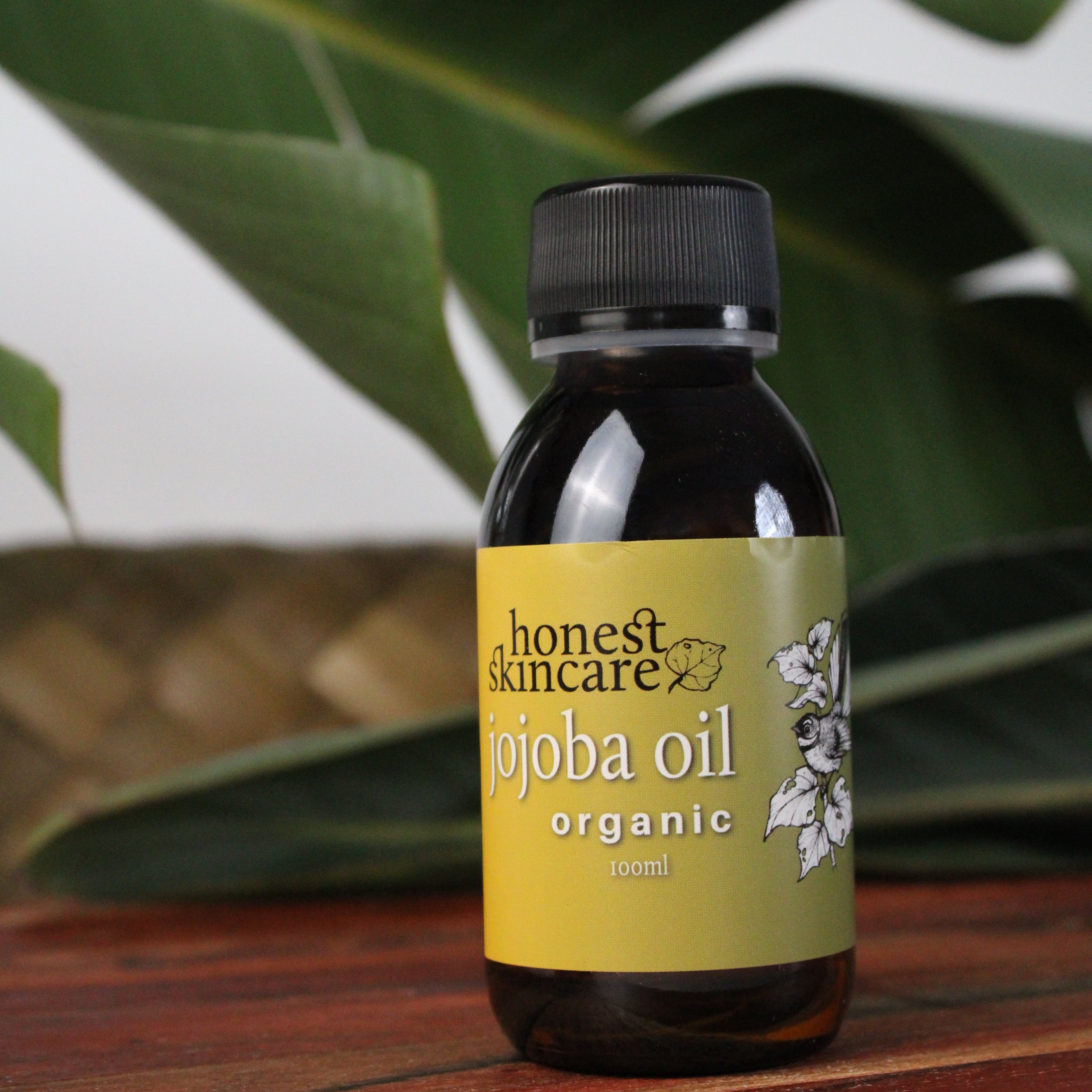 Bottle of Honest Skincare jojoba oil on a wooden surface with green leaves in the background