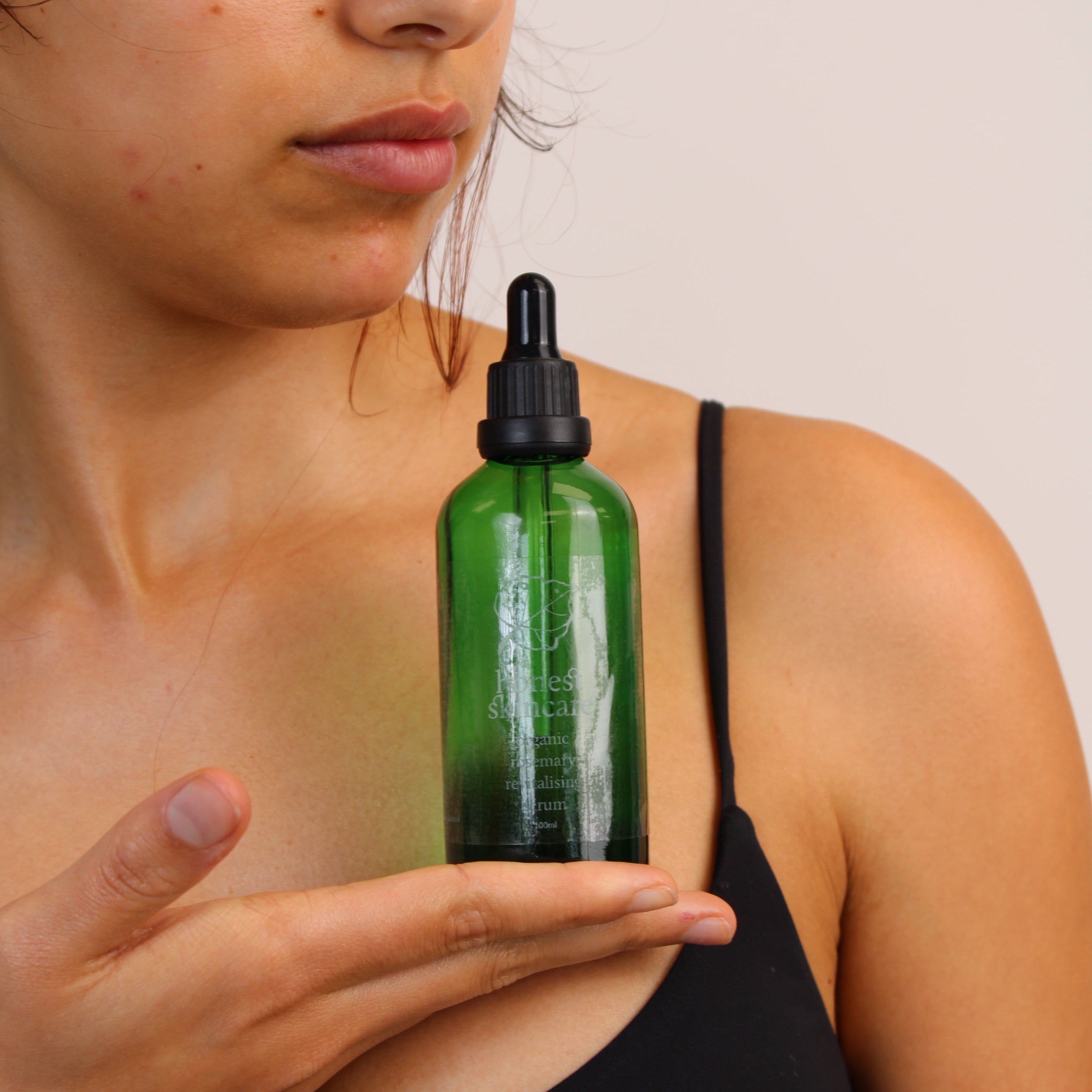 Person holding a green organic rosemary revitalising serum skincare bottle with a dropper against a neutral background