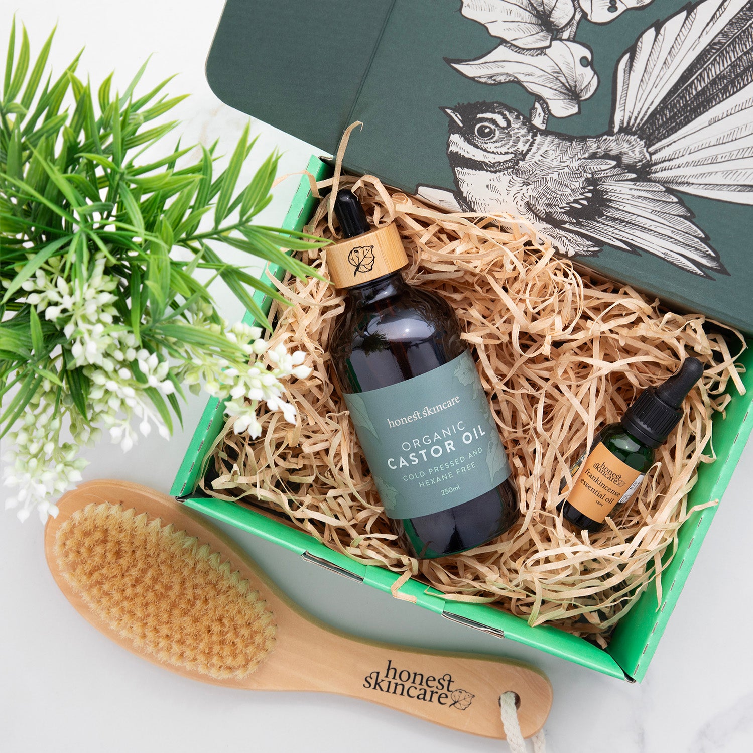 Two bottles of organic castor oil and a wooden brush in a decorative box with a plant and decorative card in the background.