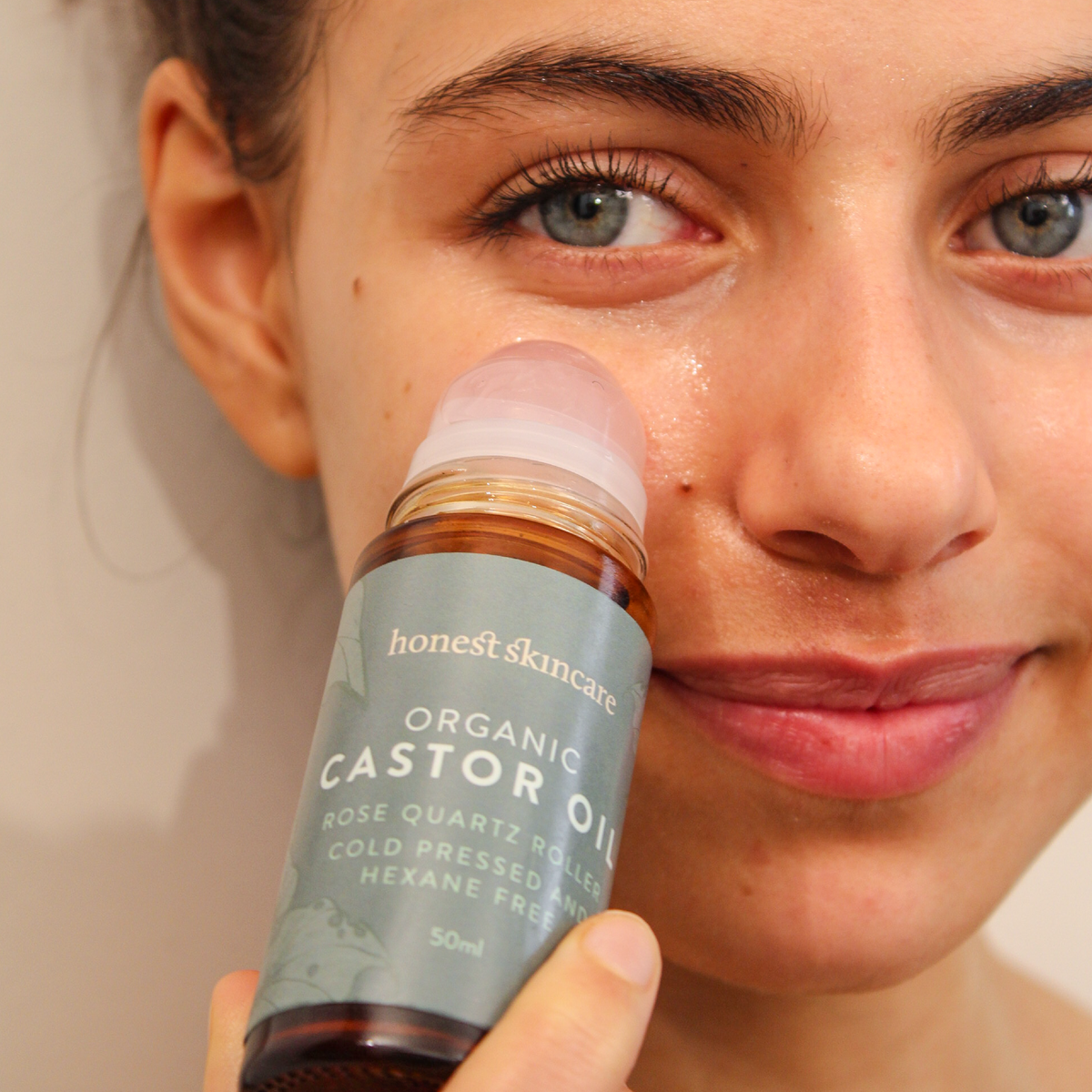 A close-up of a woman holding a bottle of Honest Skincare Organic Castor Oil next to her face, showing the product being used.