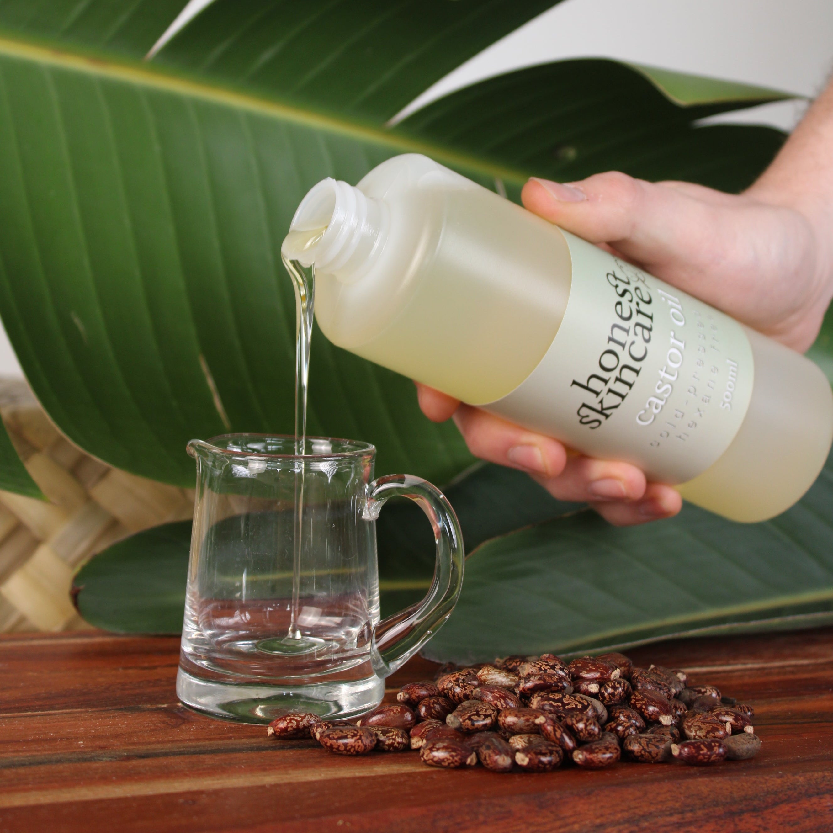 Hand holding a bottle of 'honest skincare castor oil' skincare product with a glass mug and plant in the background