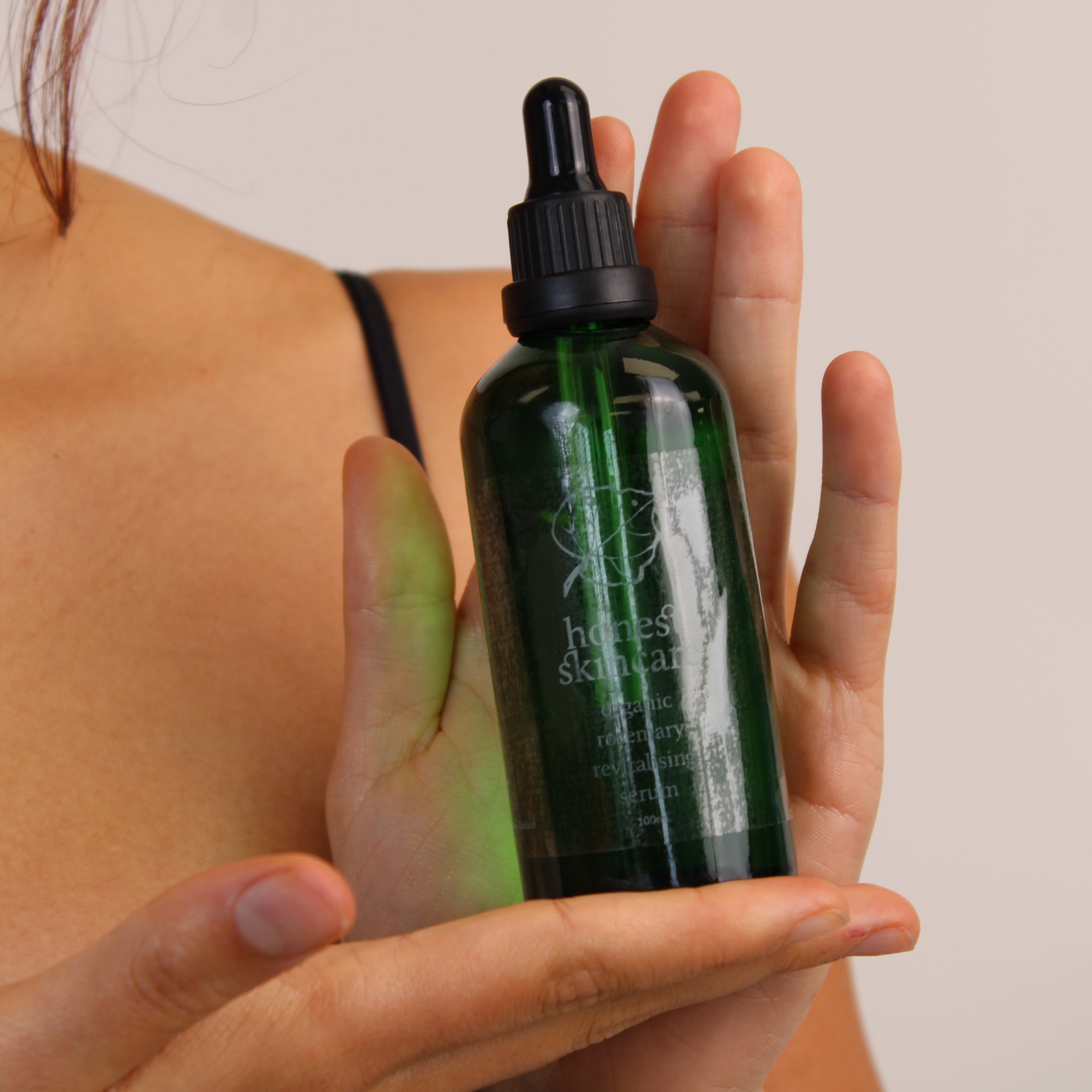 Person holding a green organic rosemary revitalising serum skincare bottle with a dropper against a neutral background