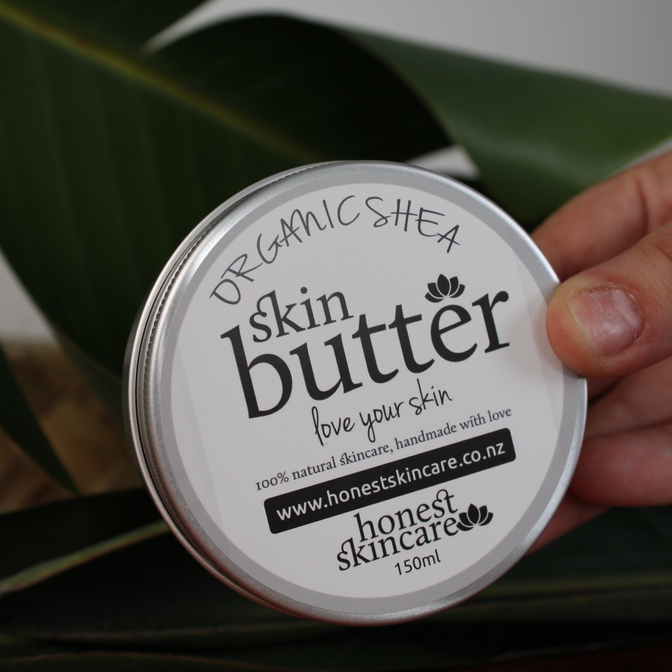 Hand holding a container of Organic shea butter tin honest skincare with leaves in the background